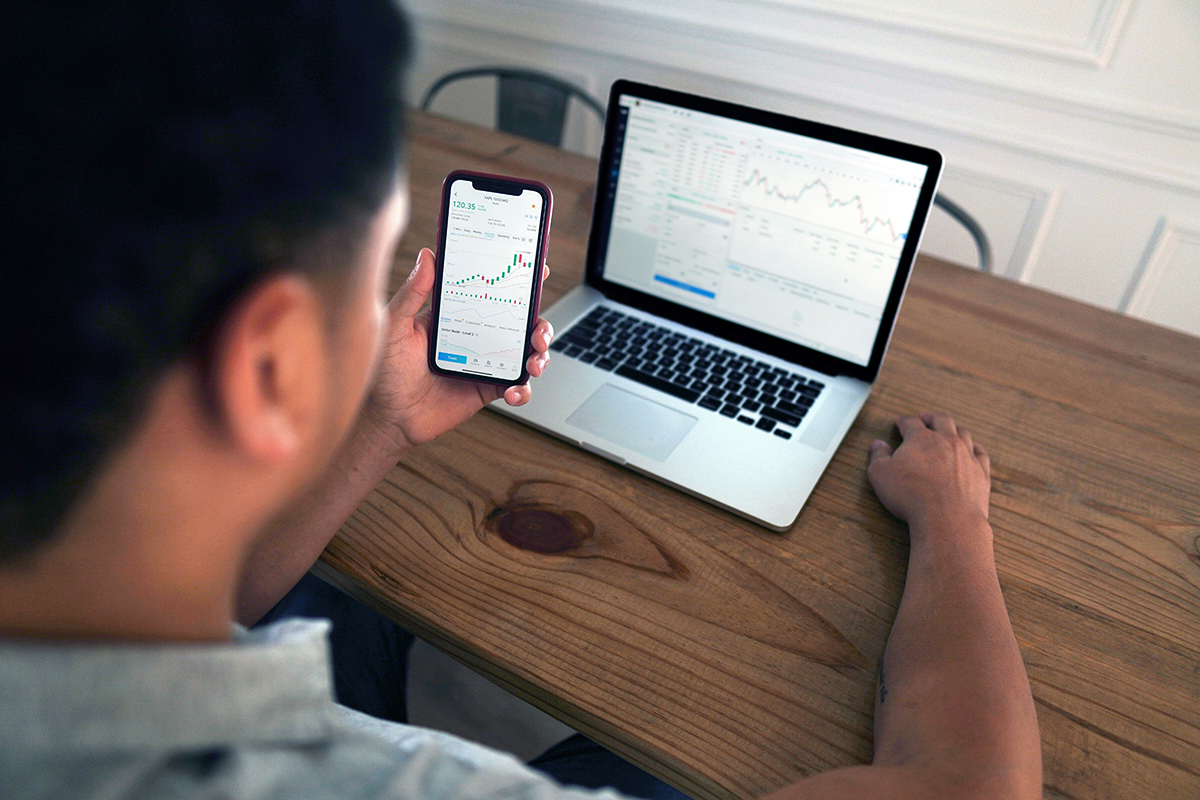 A man sat at a laptop on a desk. He has a phone in his hand. Both laptop and phone have stocks and shares graphs on them