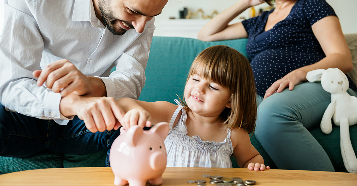 Man and woman sat on sofa with little girl putting money in piggy bank
