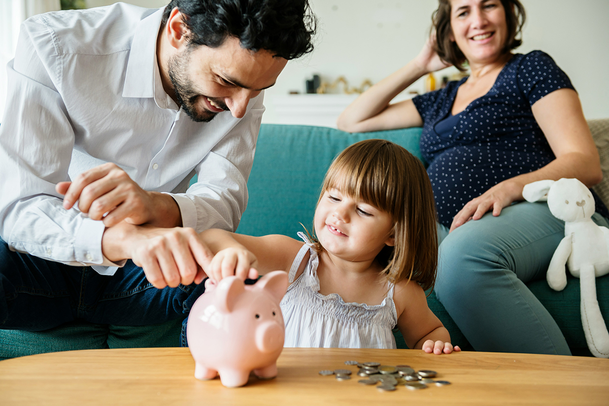 A dad sat on sofa with mum and little girl on floor putting money in piggy bank