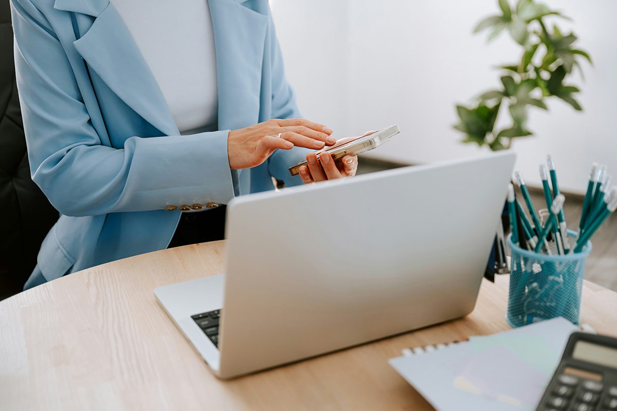 Woman sat at desk with open laptop, tapping on mobile phone