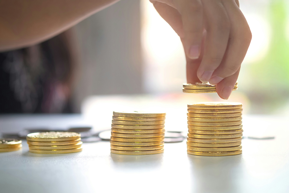 Increasing size piles of money with a hand putting additional coins on the highest pile