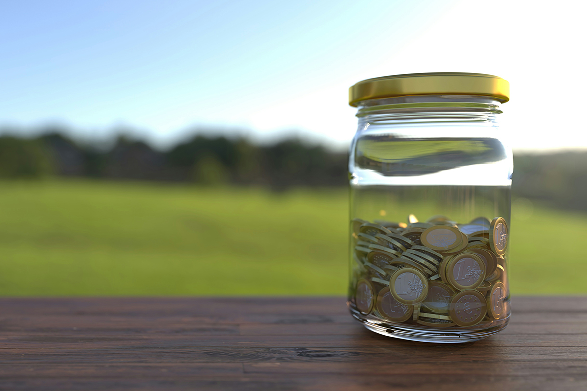 A half full jar of coins on a wooden surface with a blue sky and green grass behind