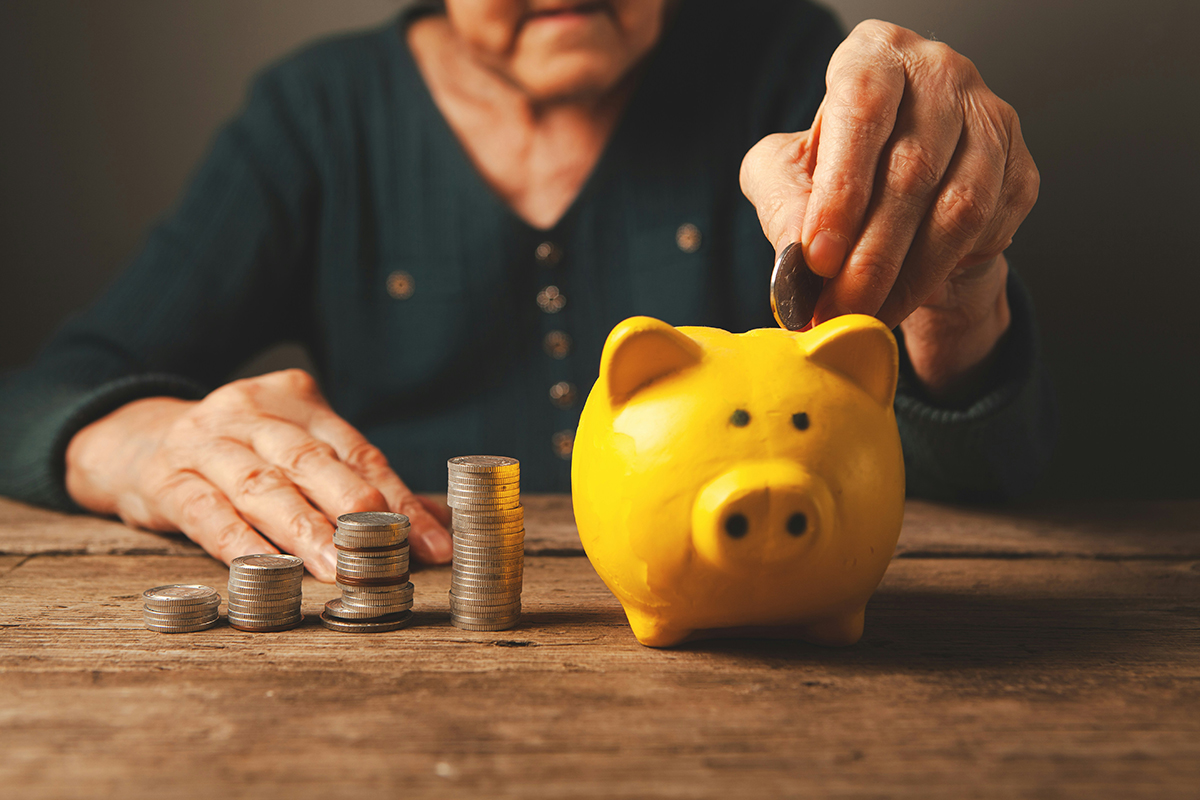 An older person putting coins in a yellow piggy bank with stacks of coins next to it